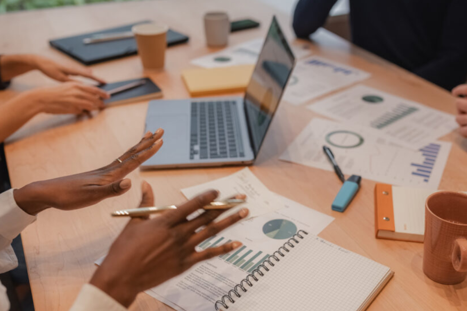 A photo showing people sitting at a table with notebooks, computers, and papers. A person is gesturing with their hands while holding a pen.