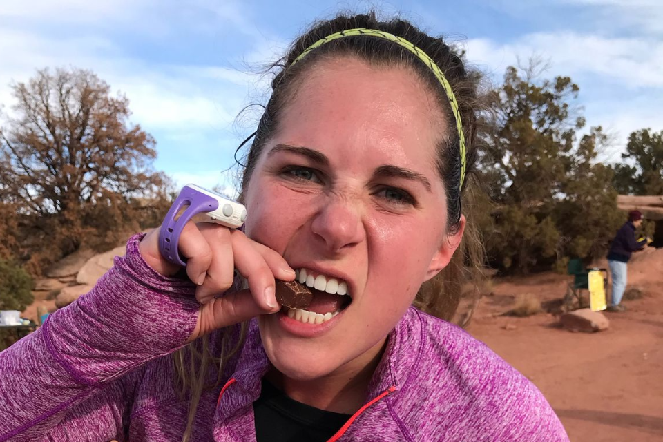 woman eating chocolate during a trail running race. holding a wrist watch. Trees behind her.