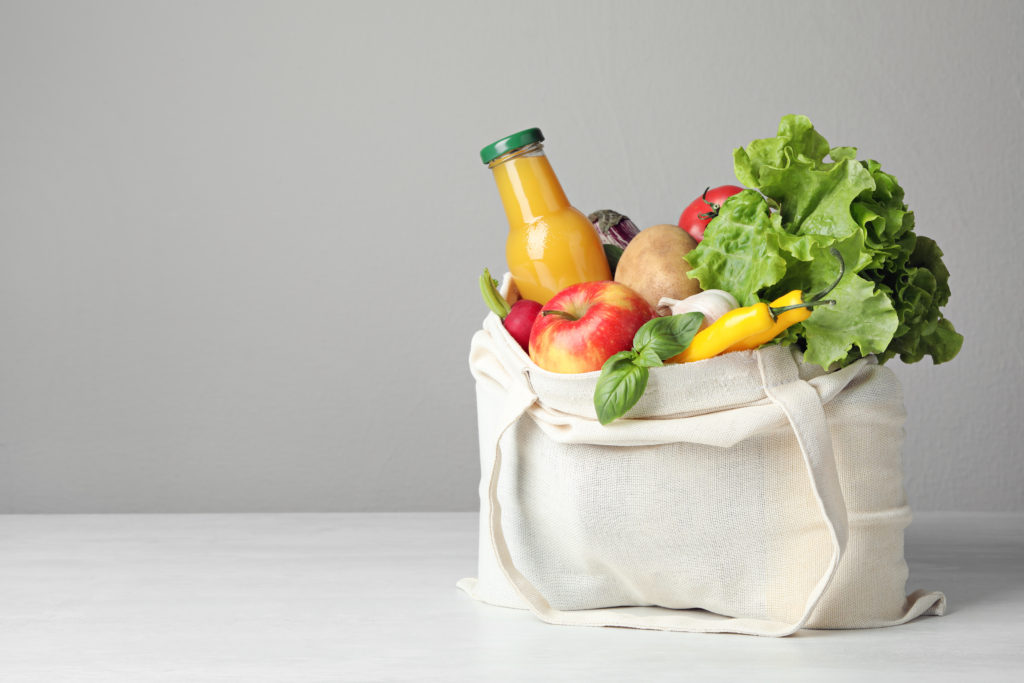 Cloth bag with vegetables and bottle of juice on table against grey background. Space for text