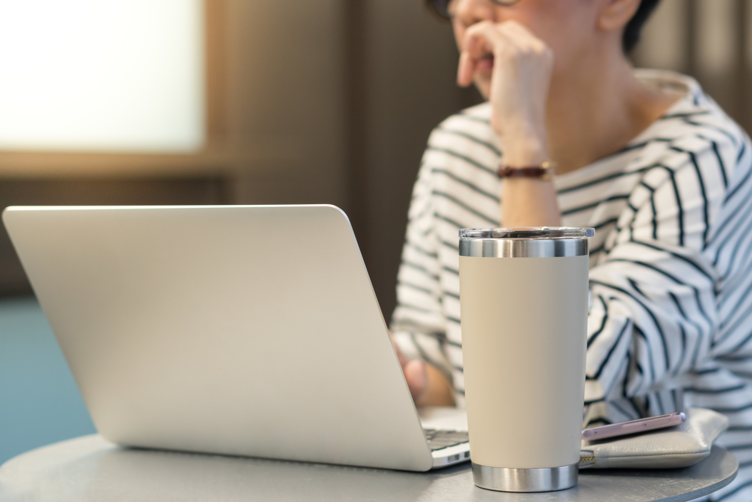 Smart looking woman use laptop computer to work from home due to Covid-19 pandemic, city lockdown and social distancing with her drink in a reusable stainless steel tumbler mug aside.