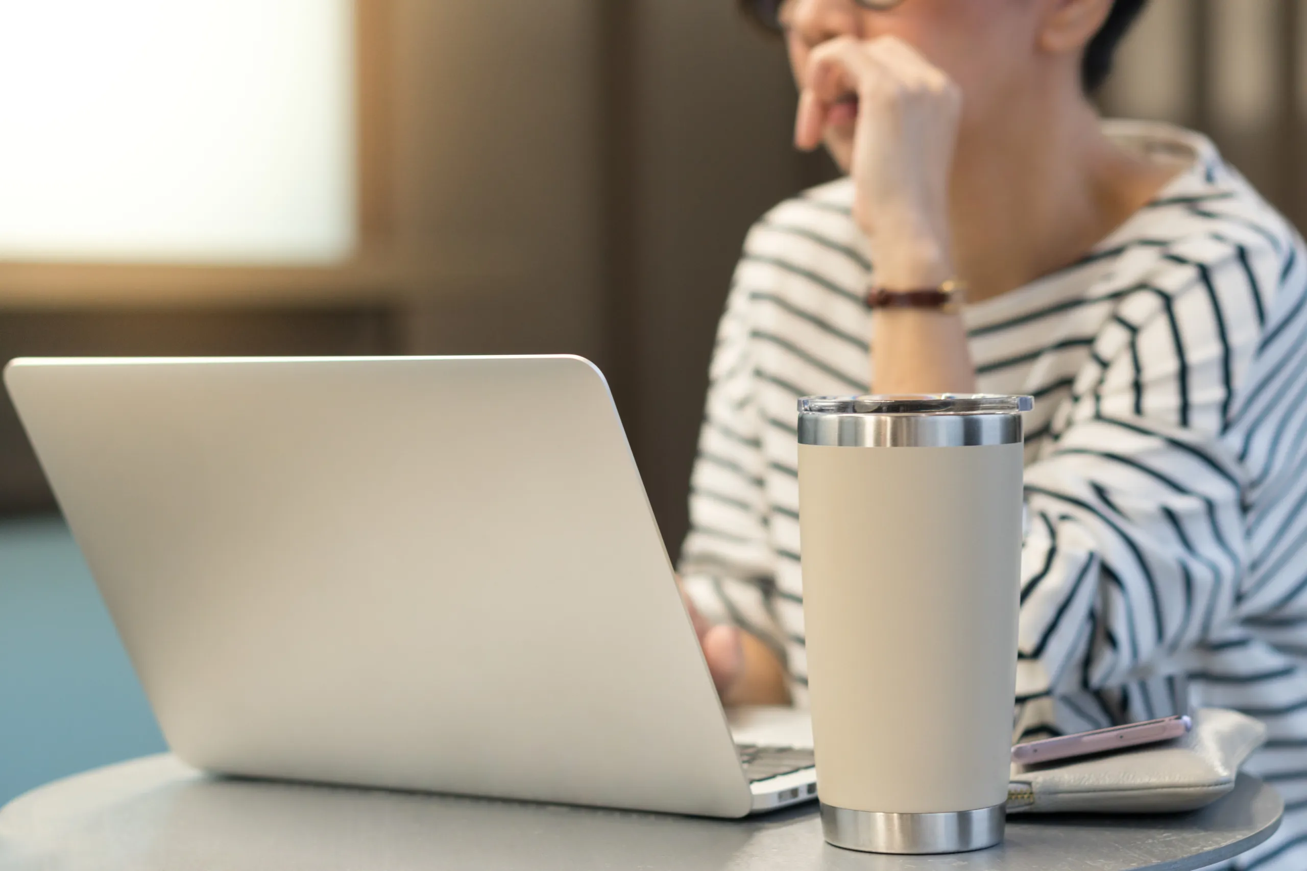 Smart looking woman use laptop computer to work from home due to Covid-19 pandemic, city lockdown and social distancing with her drink in a reusable stainless steel tumbler mug aside.