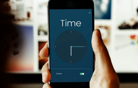 Foto de uma pessoa segurando um celular, é possível ver apenas sua mão e a tela do celular, na tela está do aparelho está um relógio, indicando o banco de horas e o pagamento de banco de horas.