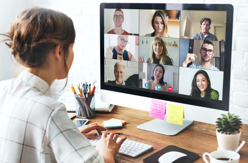 Mulher, de costas para imagem, sentada de frente para uma mesa, onde está um monitor, nesse monitor aparecem vários rostos, indicando que ela está em uma reunião. Na imagem também é possível ver suas mãos sobre um teclado, ao lado dele está posicionado um mouse.