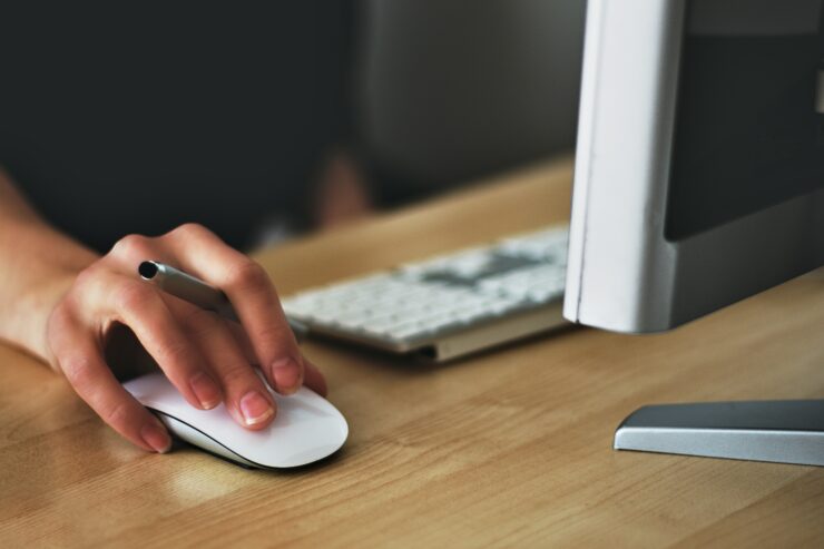 mão segurando um mouse, ao seu lado tem um teclado sob mesa de maneira e em frente um monitor