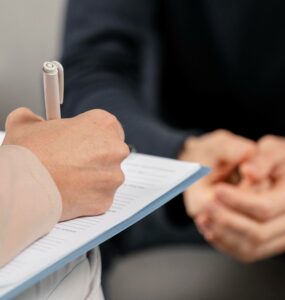 Uma pessoa está realizando anotações em sua prancheta, enquanto outra, à sua frente, responde suas perguntas, representando a entrevista que ocorre no Eneagrama da personalidade