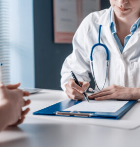 Médica sentada em sua mesa anotando as resposta de um paciente que está sentado a sua frente durante um exame