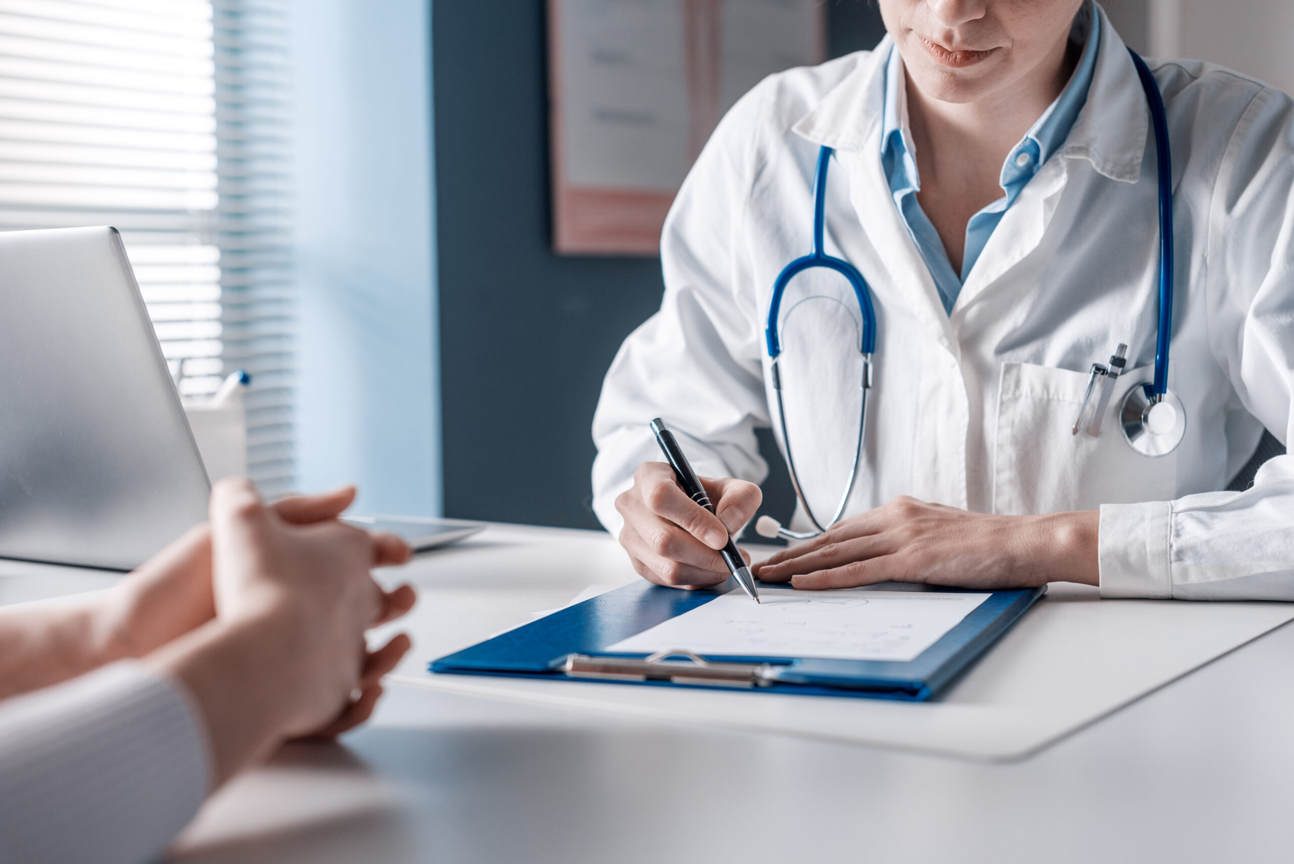 Médica sentada em sua mesa anotando as resposta de um paciente que está sentado a sua frente durante um exame