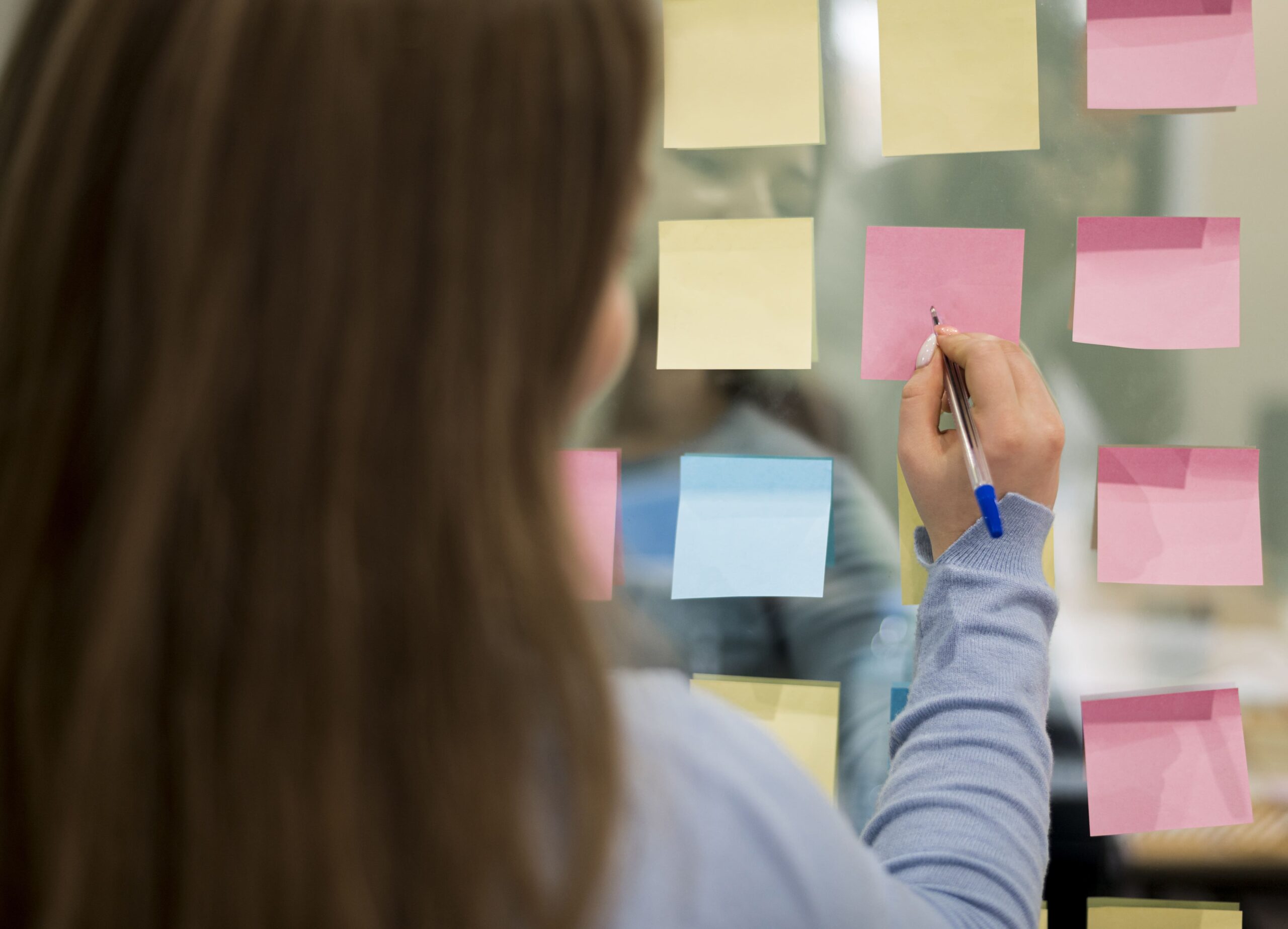 Mulher em ambiente de trabalho preenchendo pequenos papeis em um quadro, representando a organização e uma das tarefas do Product Owner na hora de pensar em suas estratégias