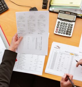 documentos e pastas sob a mesa, junto a uma calculadora, dua spessoas seguram os documentos e apontam para dados.