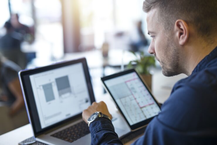 Homem em ambiente de trabalho dividindo sua atenção entre a tela de um notebook e a de um tablet.