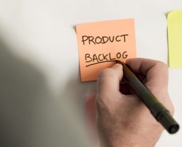 Mão escrevendo em um post it colado na parede os seguintes dizeres: Product Backlog