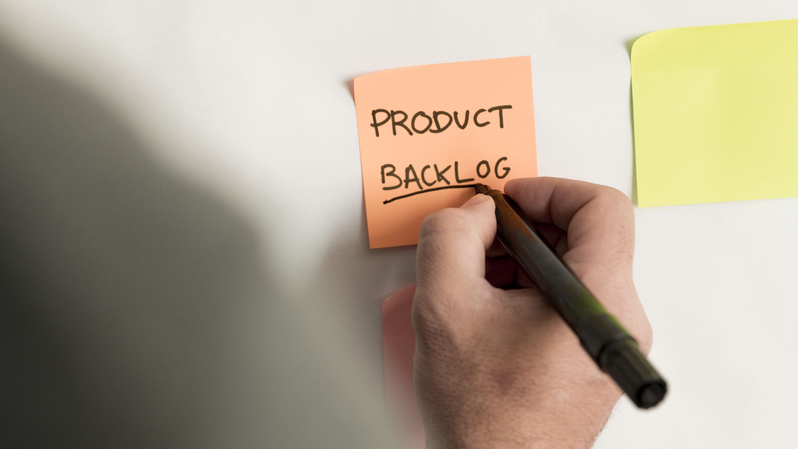 Mão escrevendo em um post it colado na parede os seguintes dizeres: Product Backlog