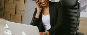 mulher sentada em frente a um notebook, com uma mãp ela digita e com a outra segura um celualr próximo a orelha e sorri