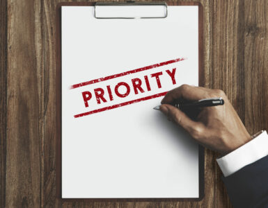 Mão com uma caneta sobre uma prancheta, onde está escrito "priority", prioridade em inglês.