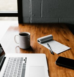 uma mesa de madeira com um notebook apoiado, aparecendo só uma aprte sob a tela, uma xícara de café, bloco em branco com uma caneta em cima e um celular em frente ao notebook