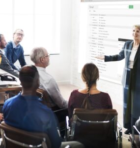 Mulher em posição de liderança falando em pé para 8 colaboradores que estão sentados à sua frente.