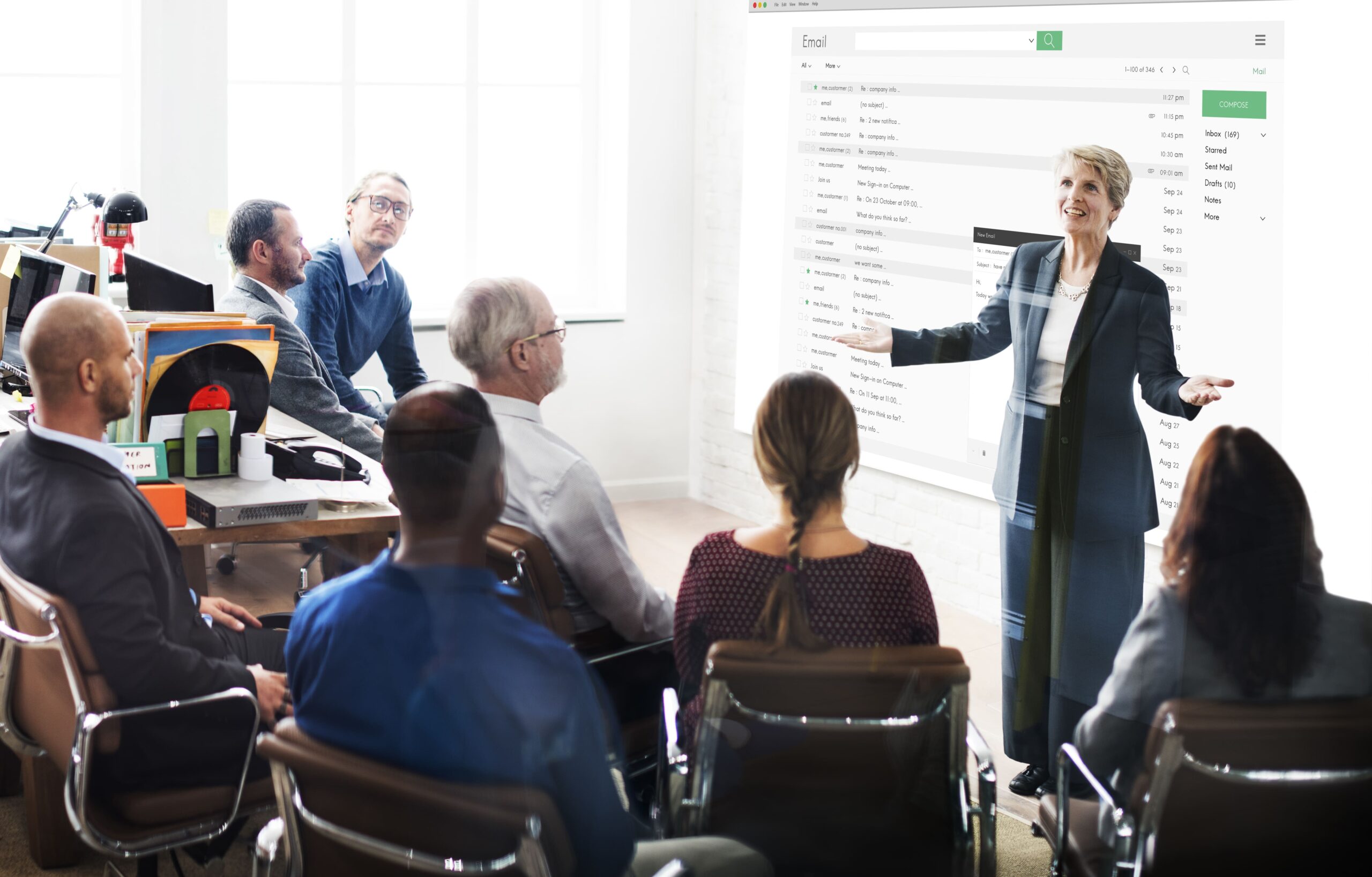 Mulher em posição de liderança falando em pé para 8 colaboradores que estão sentados à sua frente.