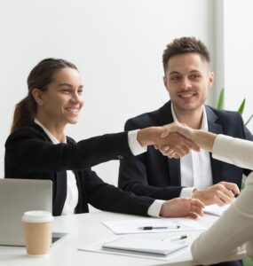 Duas mulheres se cumprimentando, sentadas cada uma em um lado da mesa, representando uma contratação, ao fundo um homem olha a cena e sorri.