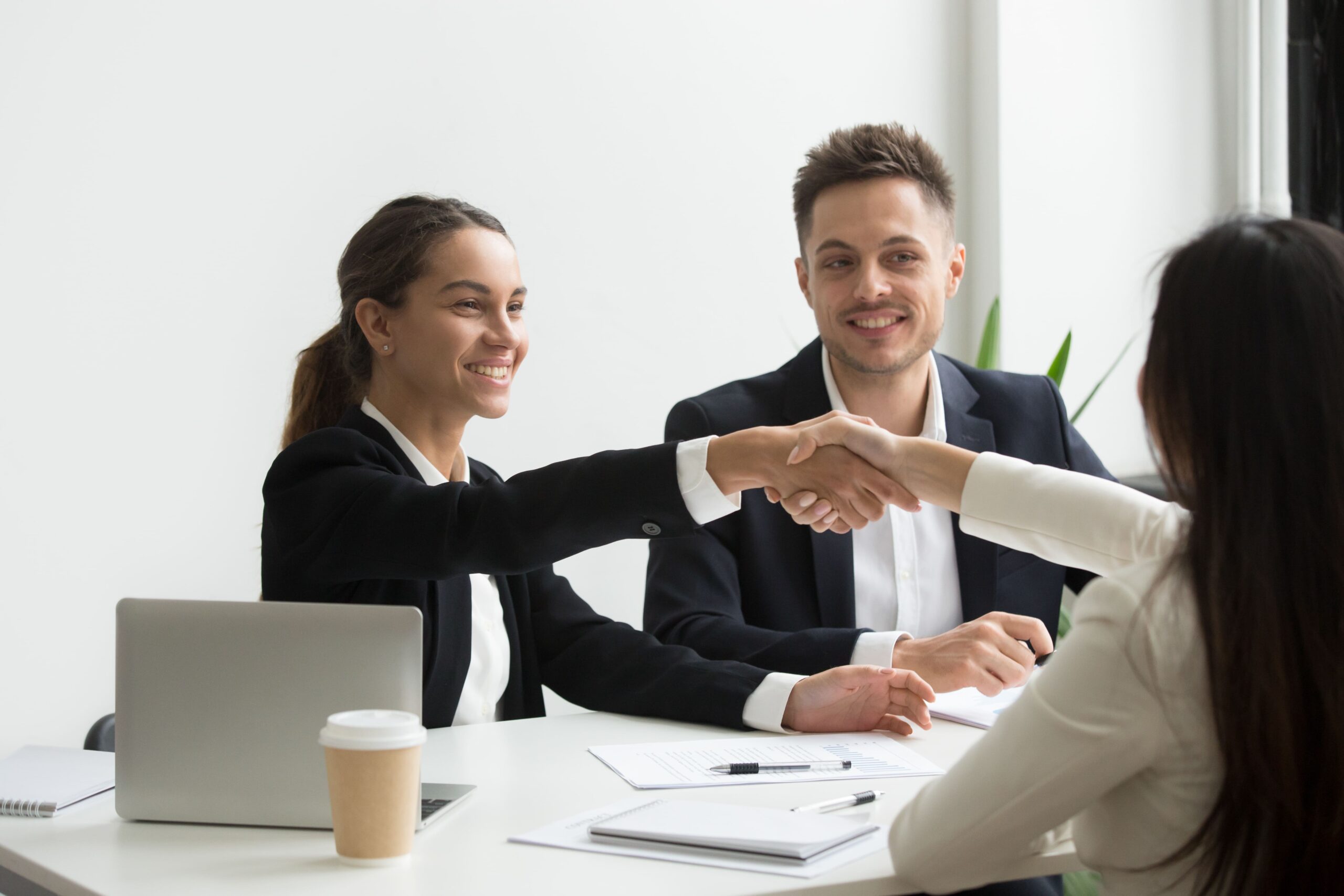 Duas mulheres se cumprimentando, sentadas cada uma em um lado da mesa, representando uma contratação, ao fundo um homem olha a cena e sorri.