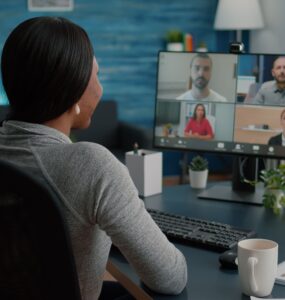 Mulher de sentada em uma cadeira de costas para a foto, a sua frente está posicionada uma mesa com um monitora em cima, em sua tela estão rostos de diferentes pessoas, representando uma vídeo conferência.