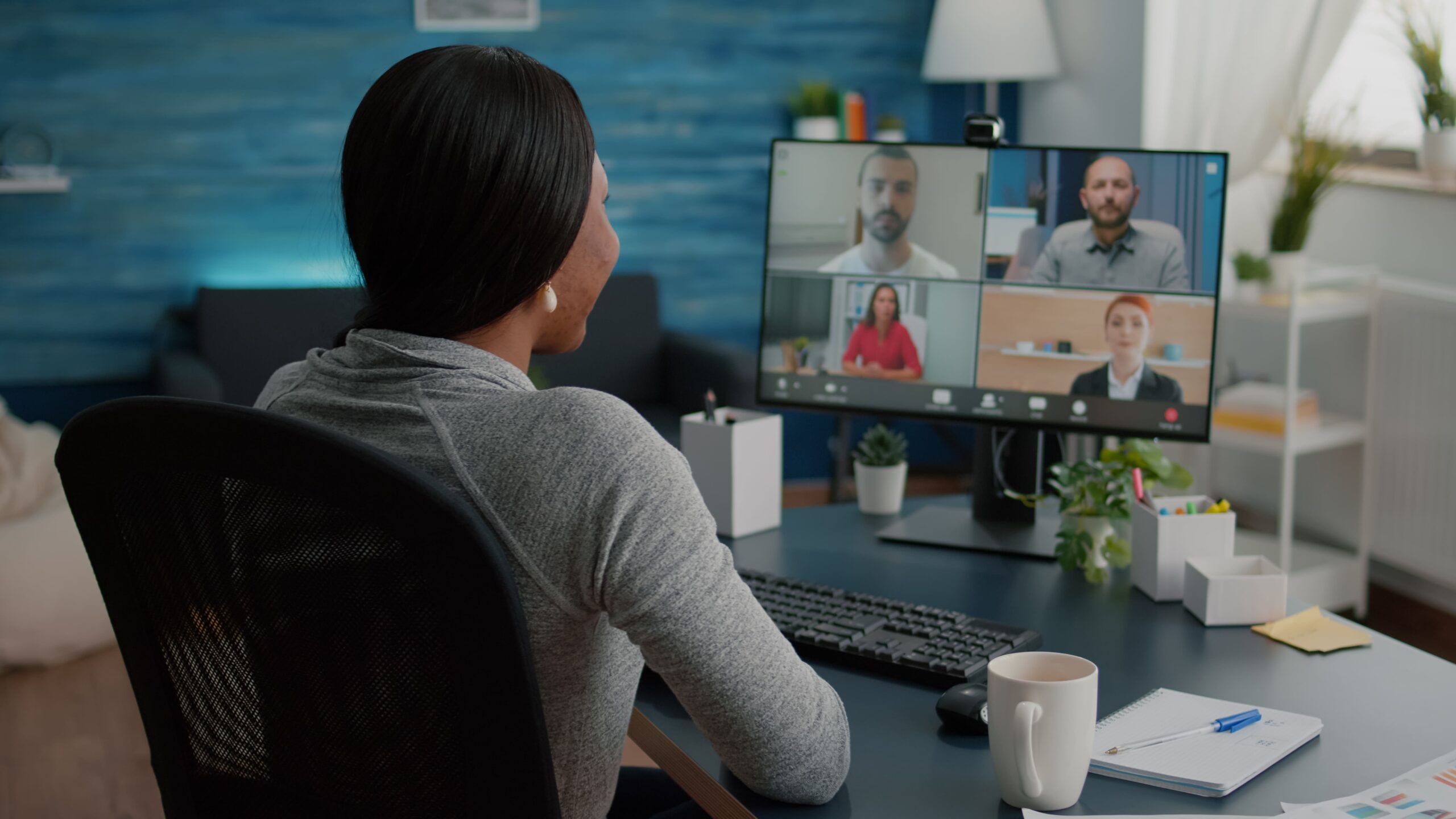 Mulher de sentada em uma cadeira de costas para a foto, a sua frente está posicionada uma mesa com um monitora em cima, em sua tela estão rostos de diferentes pessoas, representando uma vídeo conferência.