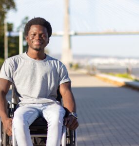 Homem negro em uma cadeira de rodas sorrindo para a foto, ao fundo uma paisagem urbana, com uma ponte.