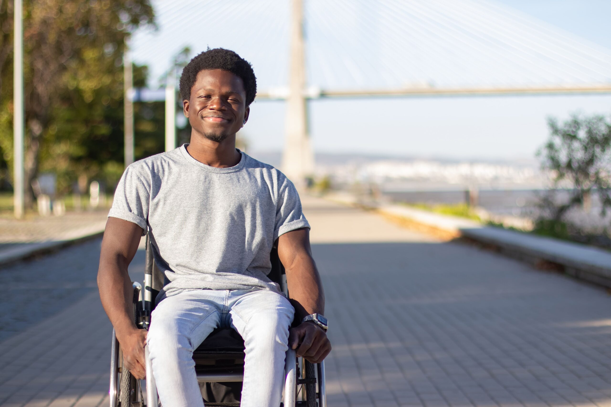 Homem negro em uma cadeira de rodas sorrindo para a foto, ao fundo uma paisagem urbana, com uma ponte.