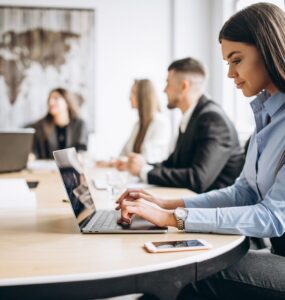 5 pessoas trabalhando em seus notebooks sentadas ao redor de uma mesa, sendo que 4 estão em segunda plano, e uma mulher está em primeiro plano.