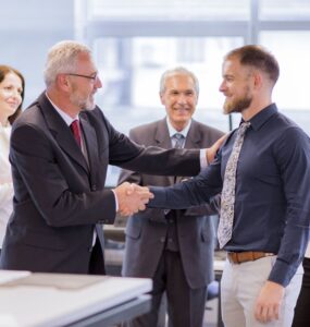 Dois homens de roupa social se cumprimentam dentro de um ambiente de escritório, ao fundo 2 mulheres e 1 homem olham a cena e sorriem.