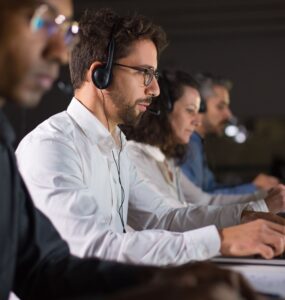 4 pessoas trabalhando uma ao lado da outra de fone de ouvido com microfone embutido e olhando para tela de seus respectivos computadores.