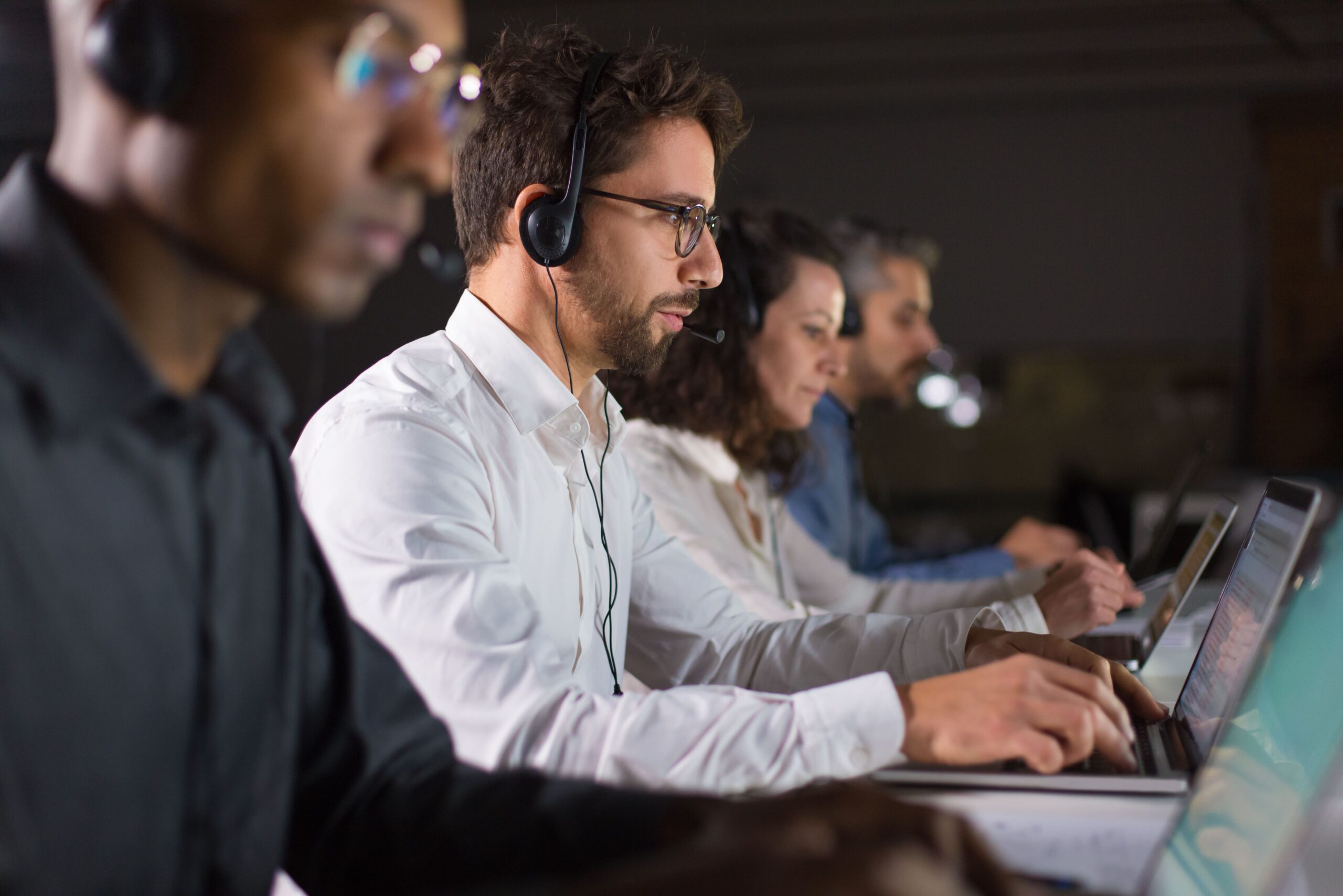 4 pessoas trabalhando uma ao lado da outra de fone de ouvido com microfone embutido e olhando para tela de seus respectivos computadores.