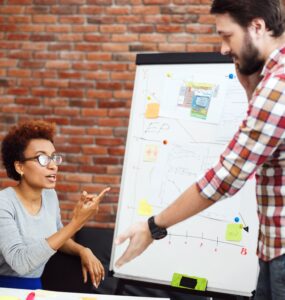 duas pessoas conversando em ambiente de trabalho em frente de um painel onde está uma apresentação.