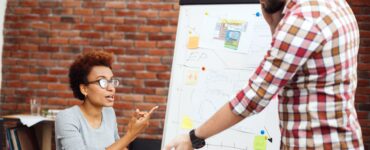 duas pessoas conversando em ambiente de trabalho em frente de um painel onde está uma apresentação.