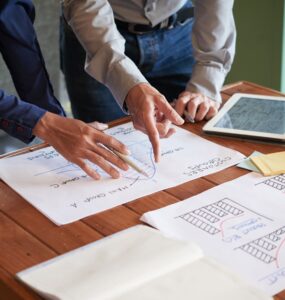 Papéis com gráficos desenhados estão em cima de uma mesa, enquanto dois homens de roupa formal apontam para eles (é possível ver apenas os seus braços e mãos), representando o planejamento de projetos.