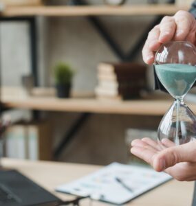 Mão de homem em roupas sociais e em ambiente de trabalho segurando uma ampulheta, ao fundo é possível ver uma mesa com um notebook em cima. A imagem representa o Time Tracking no Monday.