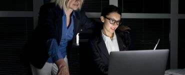 Duas mulheres em um escritório trabalhando em frente a um computador, atrás é possível ver uma janela e uma paisagem escura, representando o trabalho noturno.