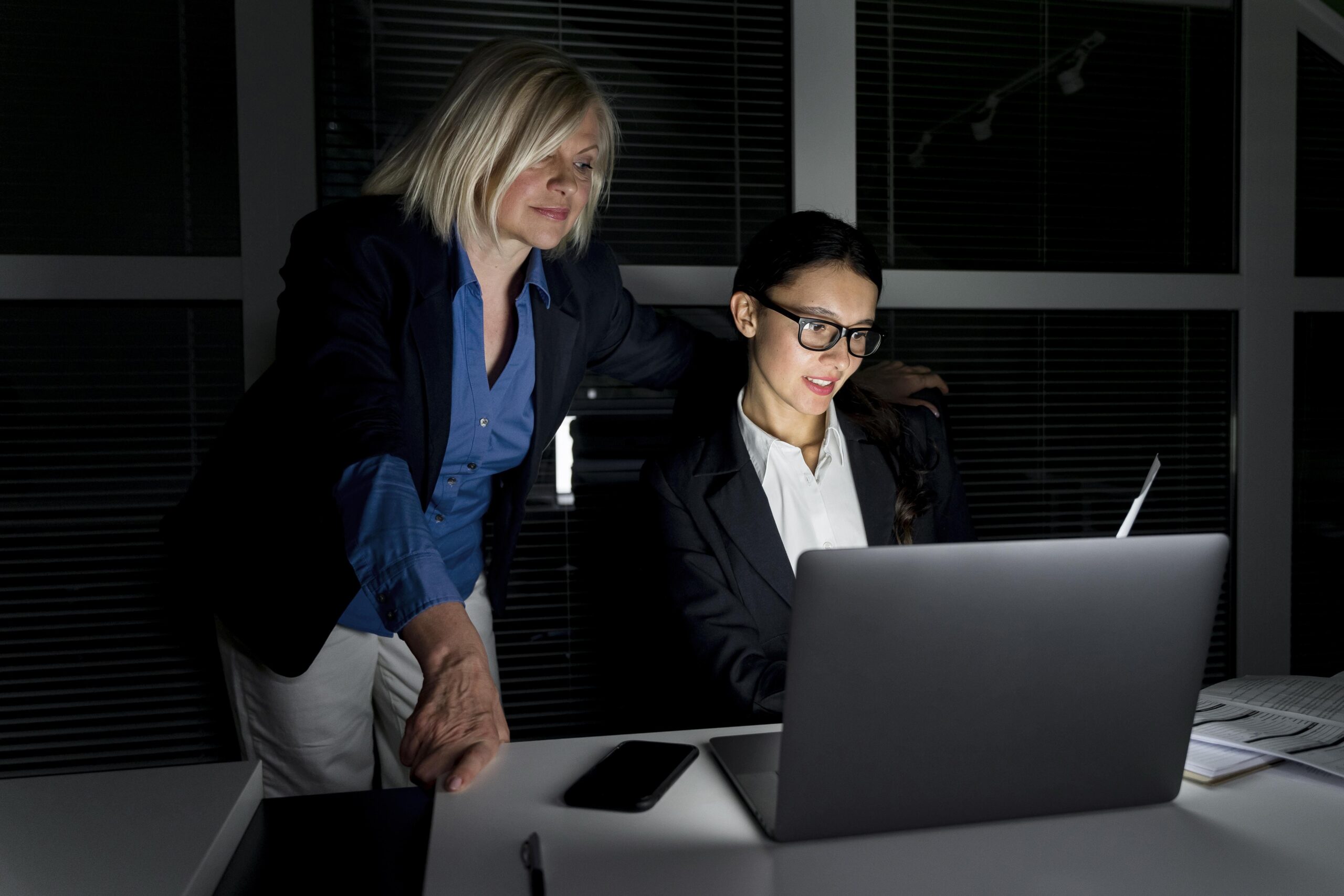 Duas mulheres em um escritório trabalhando em frente a um computador, atrás é possível ver uma janela e uma paisagem escura, representando o trabalho noturno.