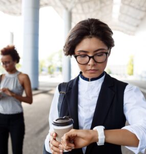 Foto de uma mulher em roupas casuais andando na rua olhando o seu relógio, e segurando na mão contraria a do relógio, ela está segurando um café. A imagem representa um trabalhador em horário de intervalo, se conectando com a hora extra intrajornada.