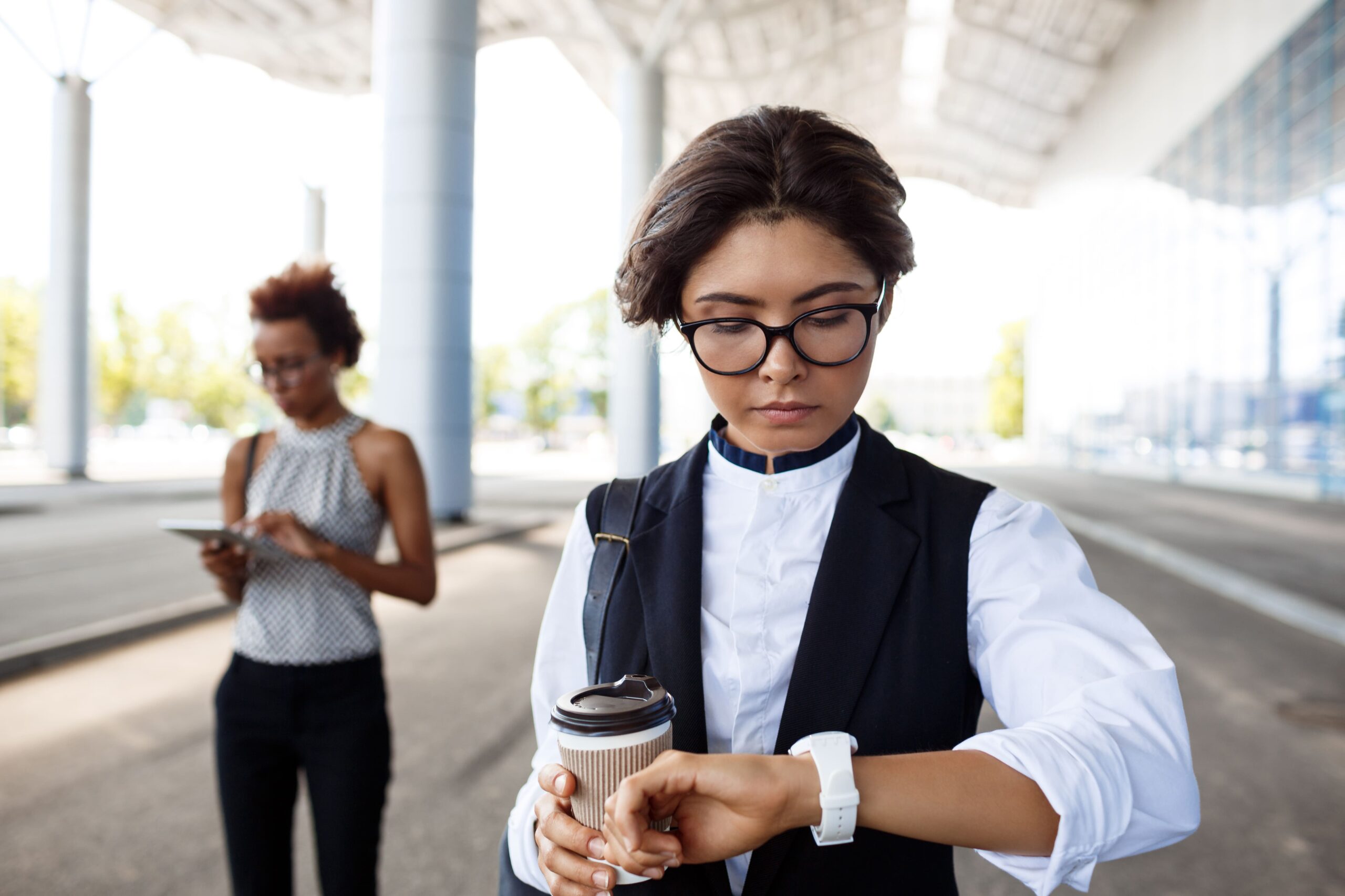 Foto de uma mulher em roupas casuais andando na rua olhando o seu relógio, e segurando na mão contraria a do relógio, ela está segurando um café. A imagem representa um trabalhador em horário de intervalo, se conectando com a hora extra intrajornada.