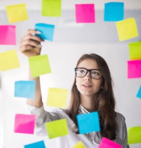 Foto de uma mulher organizando "post-its" em um vidro. A foto é tirada do outro lado do vidro, a mulher está centralizada em um buraco em meio aos "post-its". Ela está fazendo sua gestão de tarefas.