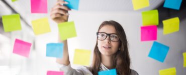 Foto de uma mulher organizando "post-its" em um vidro. A foto é tirada do outro lado do vidro, a mulher está centralizada em um buraco em meio aos "post-its". Ela está fazendo sua gestão de tarefas.