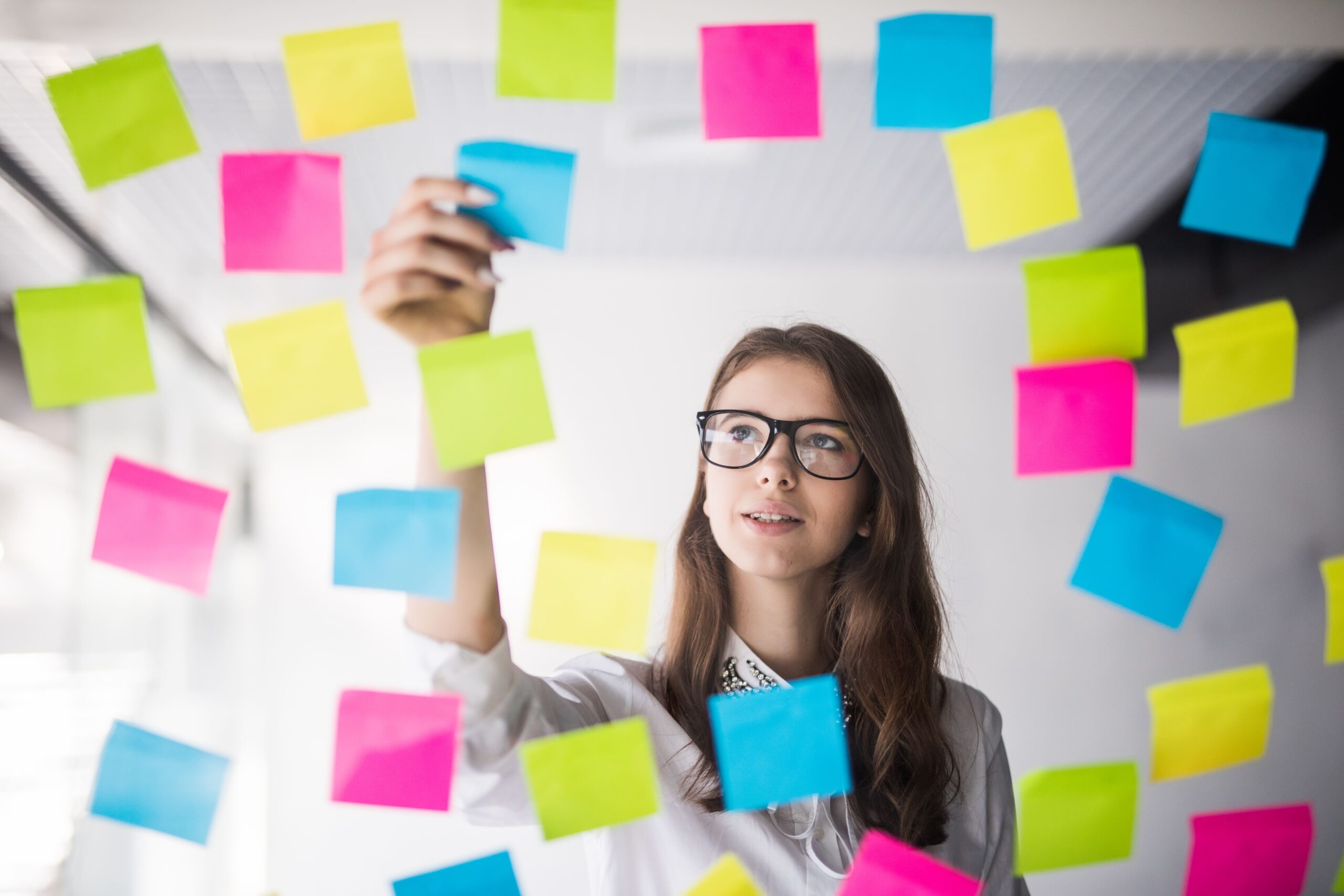 Foto de uma mulher organizando "post-its" em um vidro. A foto é tirada do outro lado do vidro, a mulher está centralizada em um buraco em meio aos "post-its". Ela está fazendo sua gestão de tarefas.