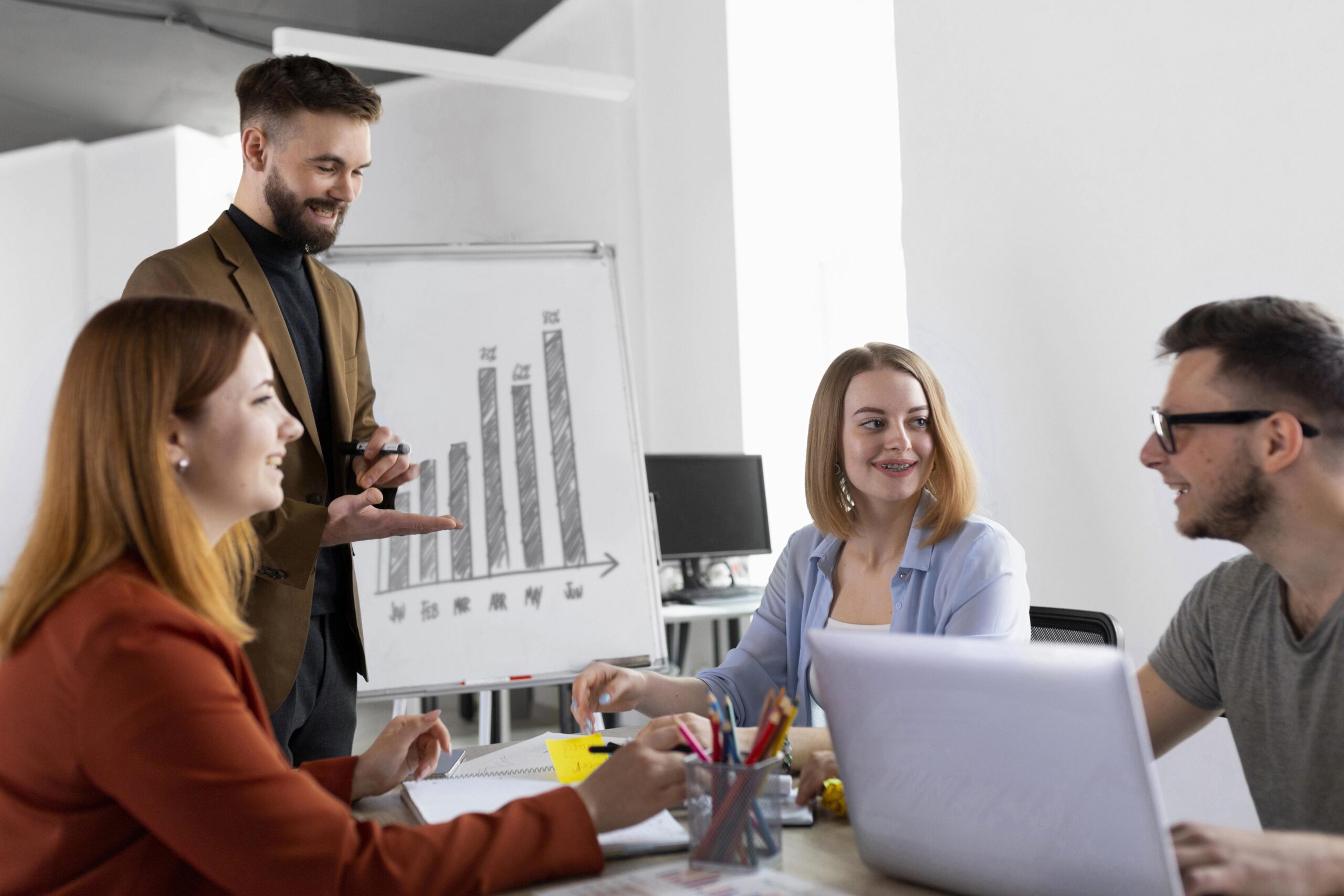 Foto de uma equipe em uma sala de reunião. Os profissionais estão interagindo e sorrindo 3 deles estão sentados em uma mesa de reunião, onde há um notebook e materiais de escritório como papeis e canetas, o outro profissional está em pé, ao fundo é possível ver um cavalete de apresentação, onde está exposto uma gráfico.