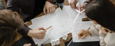 Foto de uma equipe de trabalho vista de cima. Nela e possível ver 5 pessoas de roupas sociais ao entorno de uma mesa de trabalho. Na mesa es~toa papeis onde os 5 fazem anotações e apontam. A imagem representa o processo de gestão de um projeto.