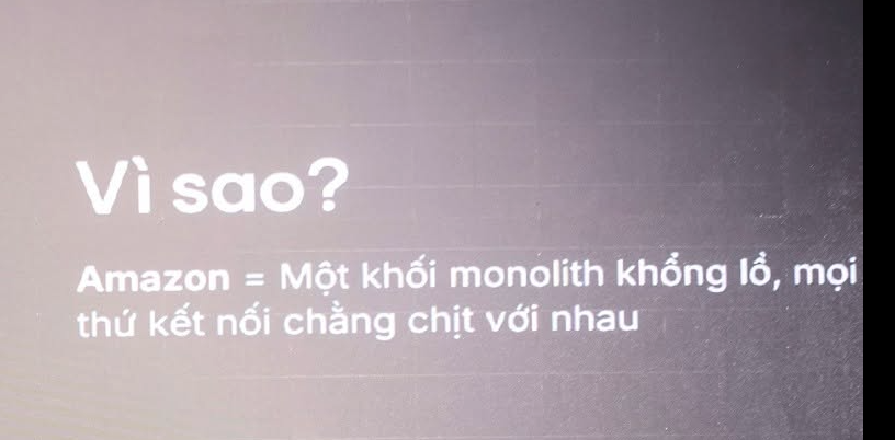 Ảnh chụp màn chiếu trong hội trường, chỉ còn phần chữ. Dòng lớn: "Vì sao?". Dòng dưới: "Amazon = Một khối monolith khổng lồ, mọi thứ kết nối chằng chịt với nhau"