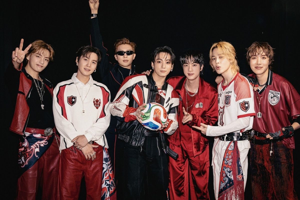 Integrantes do BTS posando com roupas vermelhas e uma bola de futebol em imagem da turnê ARIRANG