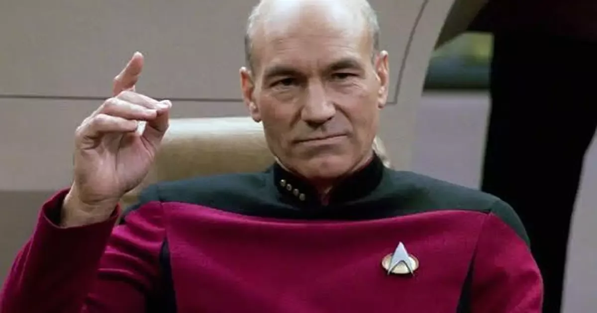 Patrick Stewart como Picard ergue a mão em um gesto de comando, vestindo o uniforme da Frota Estelar