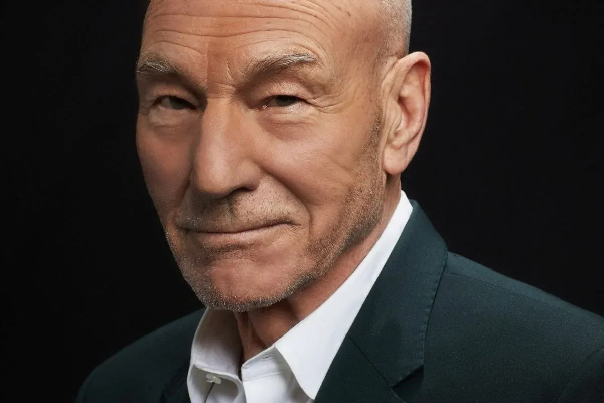 Patrick Stewart em retrato de divulgação, de terno escuro e camisa branca, olhando para a câmera com leve sorriso