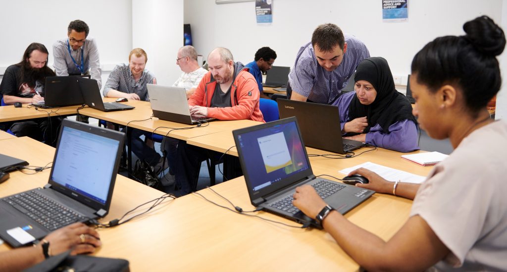 Image: Photo illustrating atmosphere of Digital Champion employment support - Several people in a training room working at laptops being showed how to use them