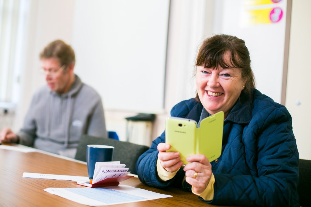 Image : A photo of a smiling woman sitting at a table with a cuppa, using her mobile phone